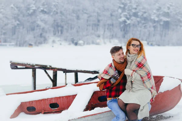 Emotional portrait of the happy couple wrapped in the knitted rug looking aside while sitting on the boat. The beautiful red head woman is sitting on the lapds of the man. — Stock Photo, Image