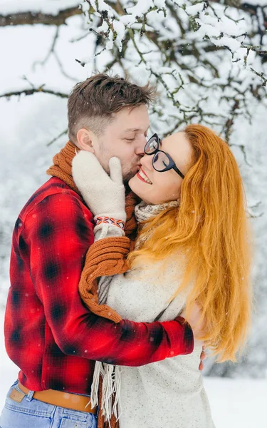 Retrato vertical de media longitud de la encantadora pareja que pasa tiempo en el bosque de invierno. El guapo novio está besando a la pelirroja en la mejilla durante la nevada. . — Foto de Stock