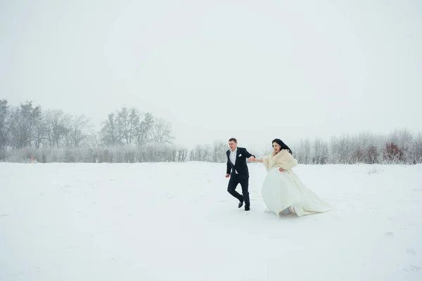 Side view of the cheerful newlywed couple holding hands and running along the snowy meadow during their walk. — Stock Photo, Image