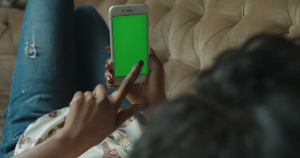 Взгляд крупным планом на афроамериканку, лежащую на диване и серфингующую в сети с помощью мобильного телефона с зеленым экраном . — стоковое видео