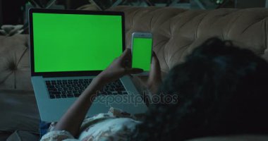 Yakın çekim arkadan görünüm Afro-Amerikan kadının bacaklarının üzerinde yeşil ekranlı dizüstü bilgisayar ile kanepede yalan ve istimal hareket eden telefon ile yeşil ekran.