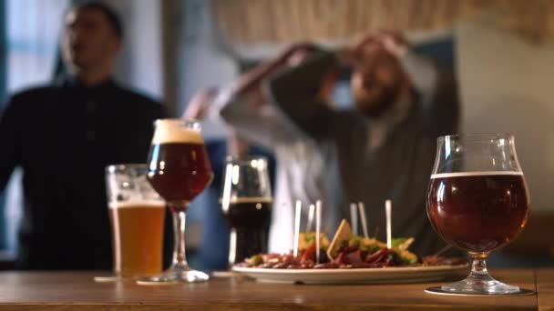 The food composition of the dark and light beer and meat plate at the blurred background of the friends cheering for their favourite football team. Side-view. — Stock Video