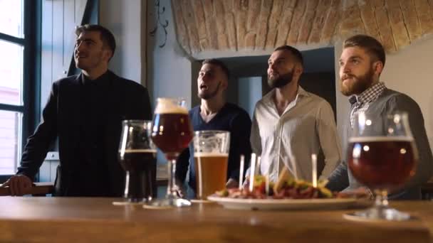 Las emociones de cuatro amigos animando a su equipo favorito. La reacción en su pérdida. La composición borrosa de la cerveza clara y oscura y el plato de carne en la barra . — Vídeos de Stock