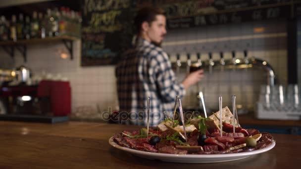 A composição do prato de carne em pé atrás do barman derramando a cerveja no copo usando a torre de cerveja rascunho e colocando o vidro na mesa . — Vídeo de Stock