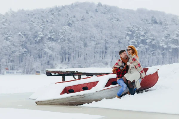 Casal alegre envolto na planície está se divertindo rindo e sentado no barco perto do rio congelado durante a queda de neve . — Fotografia de Stock