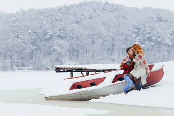 Retrato ao ar livre encantador do casal alegre beijando enquanto sentado no barco perto do rio congelado. A menina de cabelo vermelho está sentado sobre os joelhos de seu namorado . — Fotografia de Stock