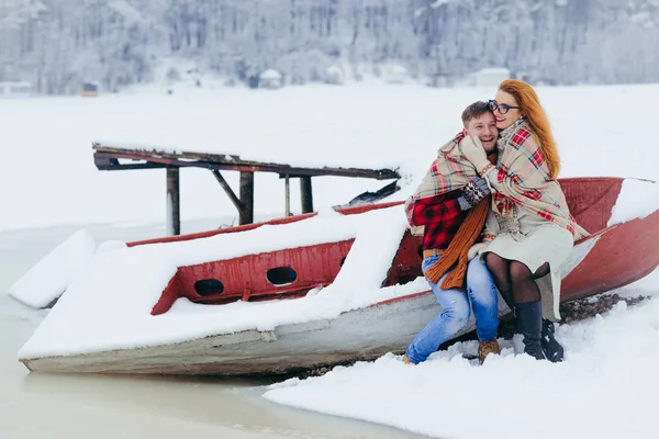 Tiro de comprimento total do belo casal amoroso envolto na planície de malha e sentado no barco perto do rio congelado durante a queda de neve . — Fotografia de Stock