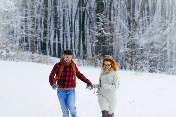 Δύο ευτυχείς εραστές κρατώντας τα χέρια και χαρωπά τρέχει κατά μήκος στο χιονισμένο δάσος, στη διάρκεια της χιονόπτωσης. — Φωτογραφία Αρχείου
