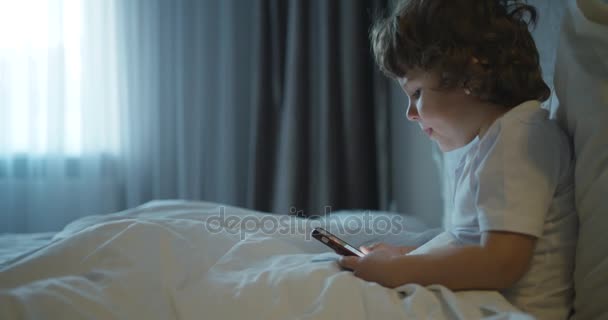 子供と技術。かわいい小さな子供は、ベッドに横たわっている間携帯電話で遊んでいます。クローズ アップの側面図です。4 k. — ストック動画
