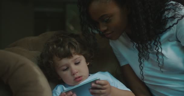 Mãe e filho estão passando um tempo juntos em casa. Pequeno filho está brincando no celular enquanto sua mãe está observando . — Vídeo de Stock