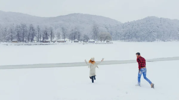 A alegre mulher cabeça vermelha está fugindo do homem jogando a bola de neve no prado nevado na floresta . — Fotografia de Stock