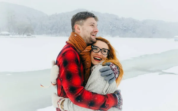 Casal amoroso alegre está rindo e abraçando durante a queda de neve no prado nevado. A bela menina cabeça vermelha com óculos . — Fotografia de Stock