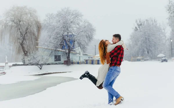 Feliz pareja amorosa se divierte en el bosque de invierno. El hombre está girando alrededor de su hermosa mujer cabeza roja durante la nevada . — Foto de Stock