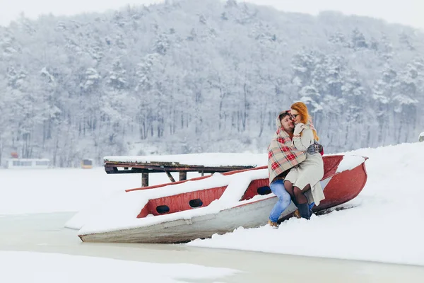 Чувствительный портрет прекрасной счастливой пары, сидящей на лодке. Красивая рыжая женщина сидит на коленях своего мужчины, завернутого в вязаную клетку во время снегопада. . — стоковое фото
