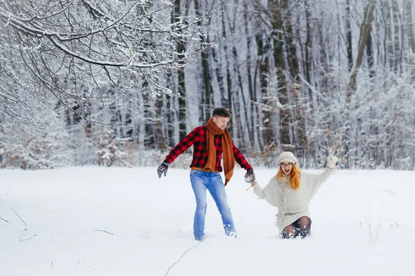 Full-length shot of the happy loving couple holding hands and running along the snowy forest. The beautiful red head woman is falling in the fluffy snow. — Stock Photo, Image