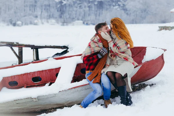 Retrato horizontal de invierno de la alegre pareja amorosa. La encantadora mujer acaricia suavemente la mejilla del hombre mientras está sentada en el barco. Están envueltos en cuadros durante la nevada. . — Foto de Stock