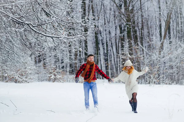 Alegre casal correndo ao longo da floresta coberta de neve durante a queda de neve . — Fotografia de Stock