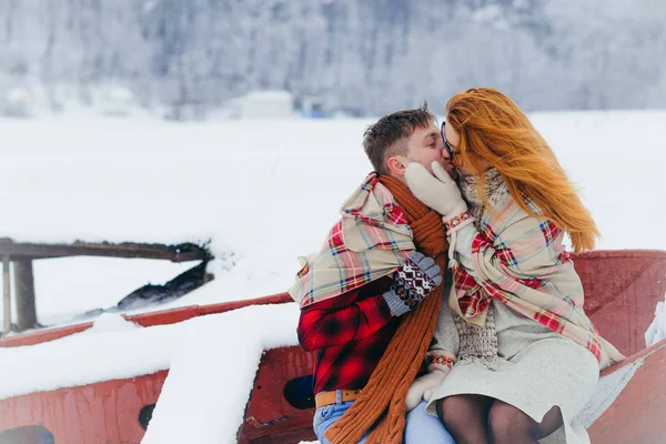 El retrato horizontal de la encantadora pareja feliz envuelta en la tela a cuadros de punto se besa mientras está sentado en el barco durante la nevada en el bosque . — Foto de Stock