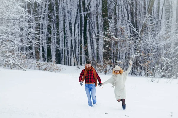 Tiro completo do casal correndo alegremente ao longo da floresta coberta de neve durante a queda de neve. Tempo de Natal. Férias de inverno . — Fotografia de Stock