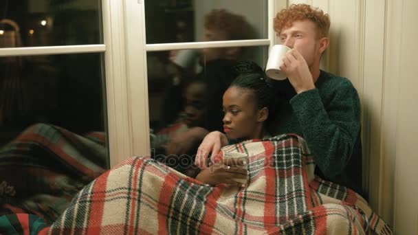 Kırmızı ekose pencere eşiğinde güzel çift sarılı. Afro-Amerikan kız beyaz Kupası ve sarışın kıvırcık saçlı adam koruyor ve sakal sıcak çay içiyor. Yatay yan portre — Stok video