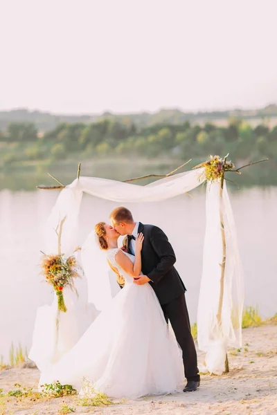 Los recién casados besos happpy durante la ceremonia de boda cerca del río . —  Fotos de Stock