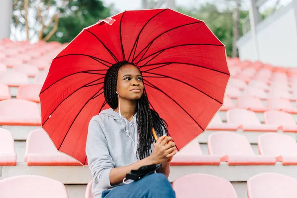 Portrait of the charming serious afro-american woman sitting under the red umbrella on the stadium.