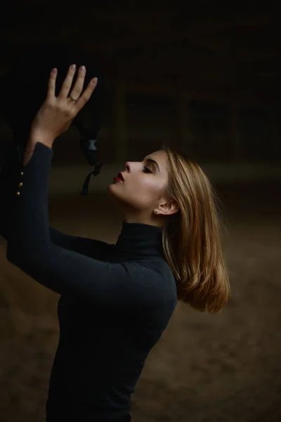 Blonde horsewoman stands in the dark hall. — Stock Photo, Image