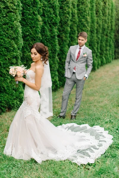 Γαμπρός στο γκρι κοστούμι φαίνεται στο νύφη που στέκεται στο γρασίδι — Φωτογραφία Αρχείου