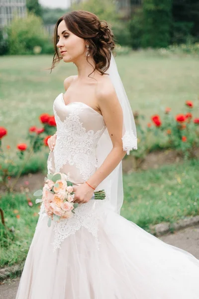 Noiva em branco vestido de renda caminha ao longo do passe com flores vermelhas — Fotografia de Stock