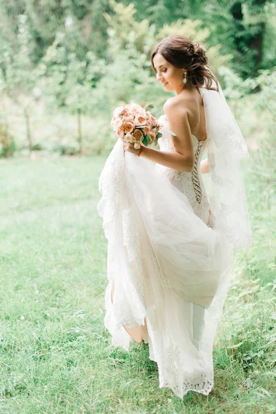 Novia sostiene vestido de novia caminando por el césped verde — Foto de Stock