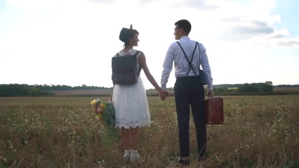 La vue arrière des jeunes mariés heureux tenant la main dans le champ ensoleillé. La mariée vêtue vintage tient le bouquet et le marié porte la vieille valise rustique . — Video