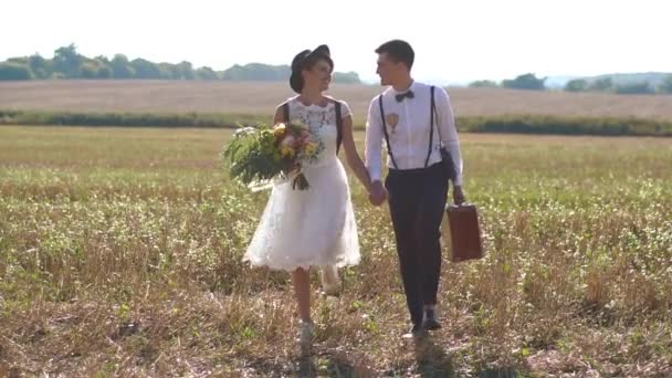 Счастливые молодожены держатся за руки и ходят по весеннему полю. Винтажная невеста держит букет желтых цветов, а жених несет старый чемодан. . — стоковое видео