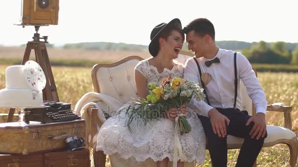 The close-up portrait of the smiling newlywed couple sitting head-to-head on the modern sofa next to the old typewriter placed on the vintage suitcases in the sunny field. — Stock Video