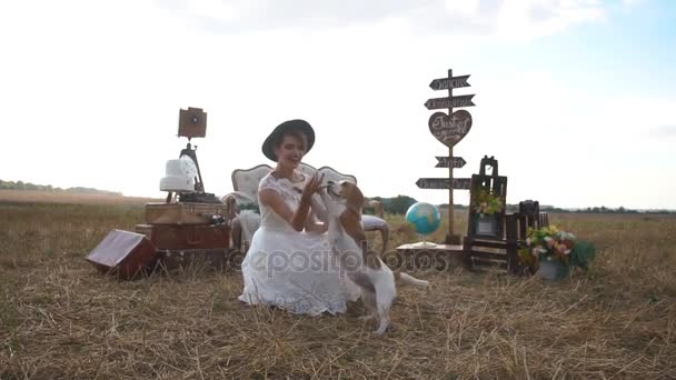 La mariée habillée vintage souriante joue avec le chien mignon à l'arrière-plan de la composition composée des vieilles valises, globe, plaques en bois et meubles rustiques . — Video