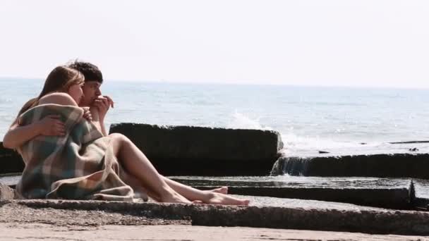 La encantadora pareja está pasando un buen rato besándose y acostándose en la playa cerca del mar. Están envueltos en el tejido a cuadros . — Vídeos de Stock