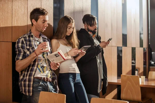 Três jovens de pé na sala perto da parede com gadgets em suas mãos . — Fotografia de Stock