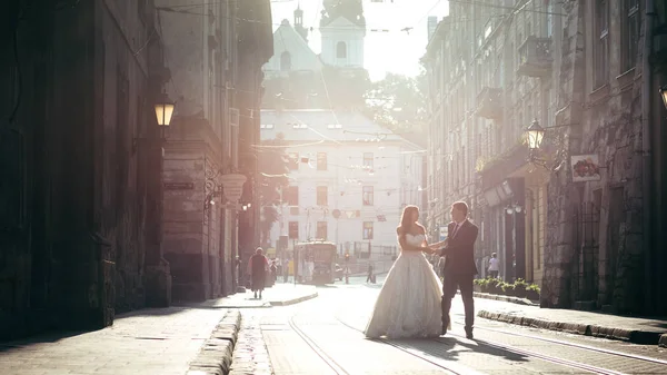 Full-length wedding shot of the stylishly dressed newlywed couple talking and holding hands during their walk along the road of the city centre. — Stock Photo, Image