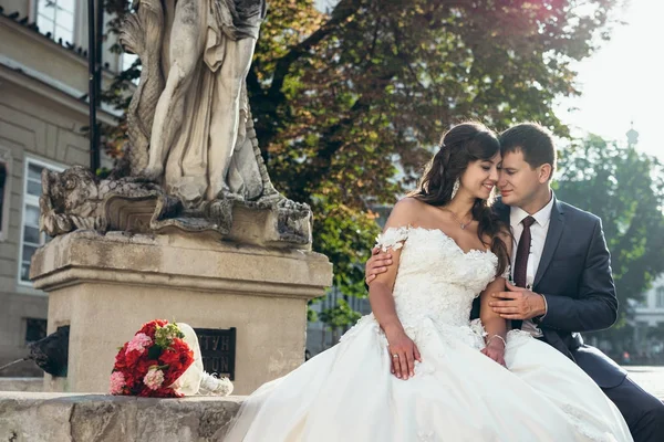 Charmiga kärleksfull bara gift par kramas och sitter på fontänerna nära bröllop bukett av röda blommor. — Stockfoto