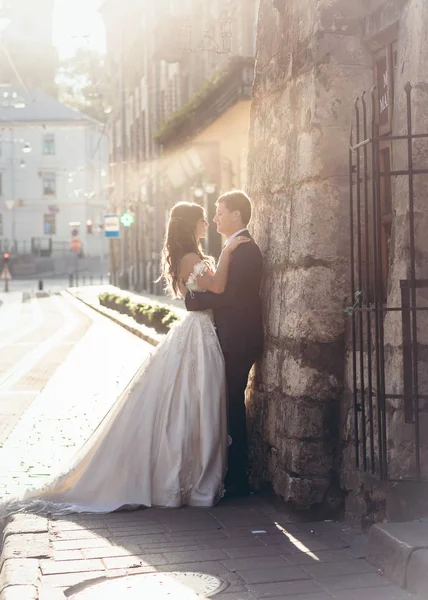 Caminhe dos recém-casados alegres ao longo da cidade velha durante o pôr do sol. Acarinhosamente abraçando belo par. O noivo está inclinado na parede. . — Fotografia de Stock