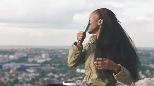 Side view of the young charming african girl brushing her long dark hair at the background of the panorama city. — Stock Video