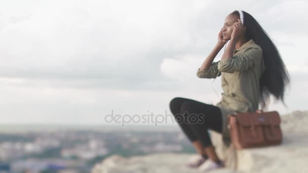 Side portrait of the charming young african girl putting on the headphones and using the mobile phone at the background of the panorama city. — Stock Video