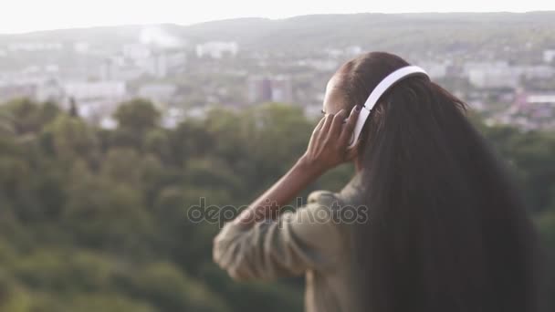 Πίσω όψη από το ελκυστικό αφρο-αμερικανικό κορίτσι με μακριά καστανά μαλλιά βάζοντας τα ακουστικά και απολαμβάνοντας τη θέα στην πόλη. — Αρχείο Βίντεο
