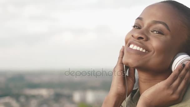 Närbild porträtt av den vackra leende afro-american flickan att sätta på hörlurarna och njuta av musiken på bakgrunden till staden panorama. — Stockvideo