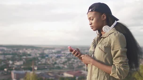 Portret van de kant van de ernstige Afrikaans meisje in de hoed SMS, chatten en surfen via de mobiele telefoon op de achtergrond van het panorama van de stad. — Stockvideo