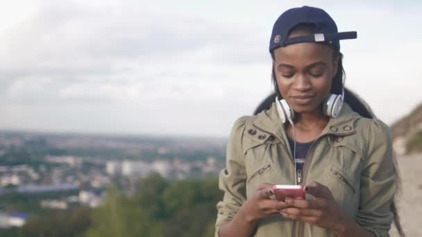 Retrato frontal de la hermosa chica africana con maquillaje natural y sonrisa encantadora está enviando mensajes de texto a través del teléfono móvil en el fondo de la ciudad panorámica . — Vídeo de stock