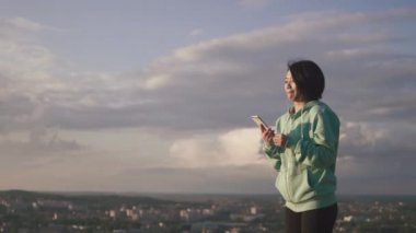 Sohbet ve panorama şehrin arka plan, cep telefonu üzerinden tarama genç çekici Asya kız kulaklık manifatura portresi.