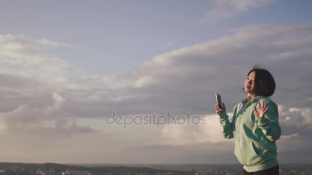 Cheerful young asian girl with lovely smile is listening to music via the earphones and dancing at the background of the sky. — Stock Video