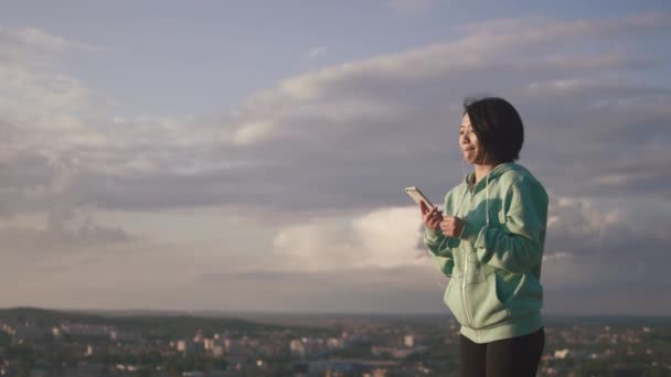 Portret van de jonge aantrekkelijke Aziatisch meisje in de oortelefoons SMS, chatten en surfen via de mobiele telefoon op de achtergrond van de panorama-stad. — Stockvideo
