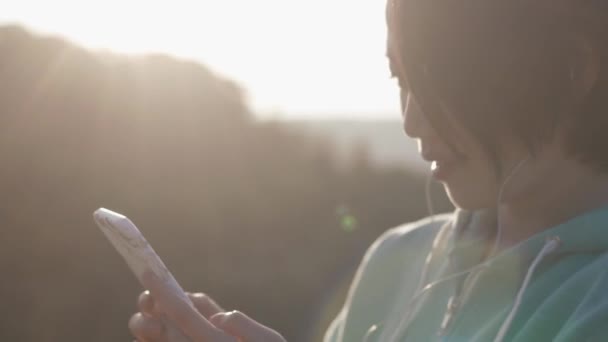 Close-up portret van de jonge Aziatische meisje met shot donker haar chatten via de mobiele telefoon in de zon. Buiten concept. — Stockvideo