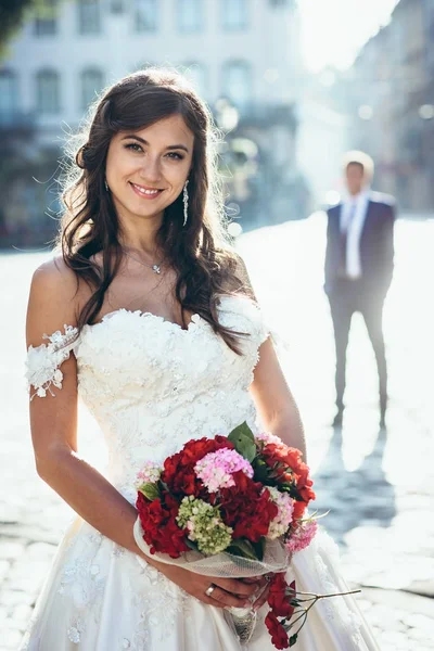 Retrato da noiva morena com maquiagem natural e sorriso encantador segurando o buquê de casamento das flores vermelhas e rosa no fundo borrado do noivo . — Fotografia de Stock